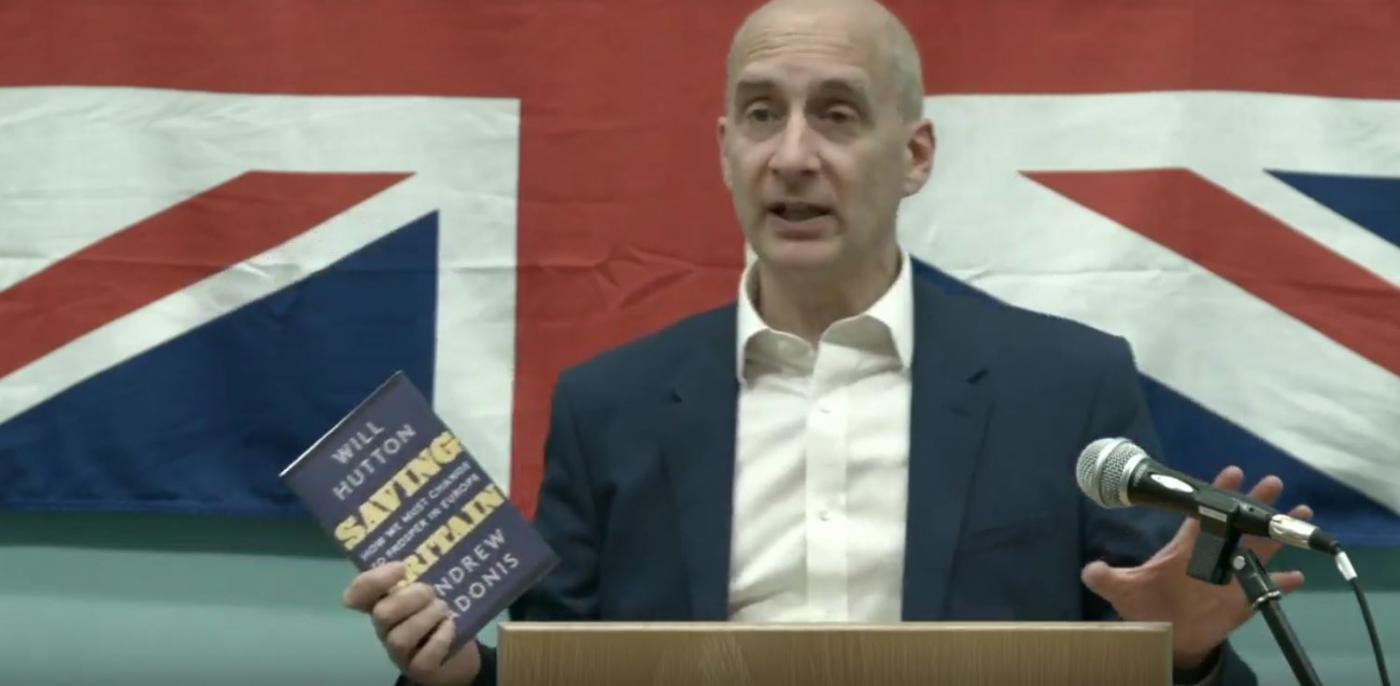 Andrew Adonis holding up copy of his book in front of Union Jack flag
